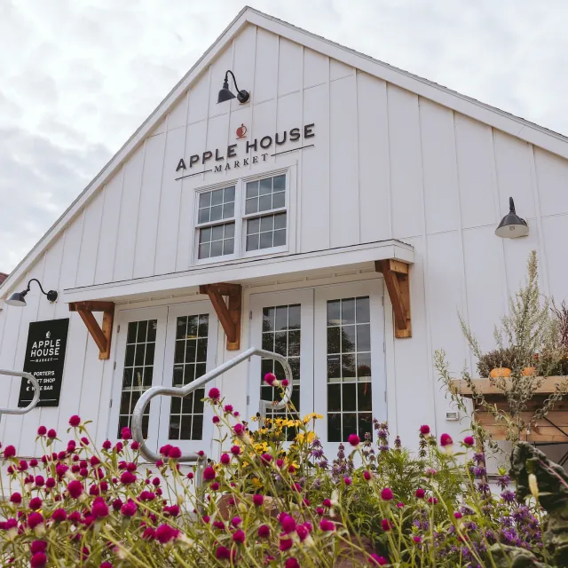 White barn-style building labeled "Apple House Market" with double doors. Colorful flowers in foreground, creating a welcoming, rustic atmosphere.