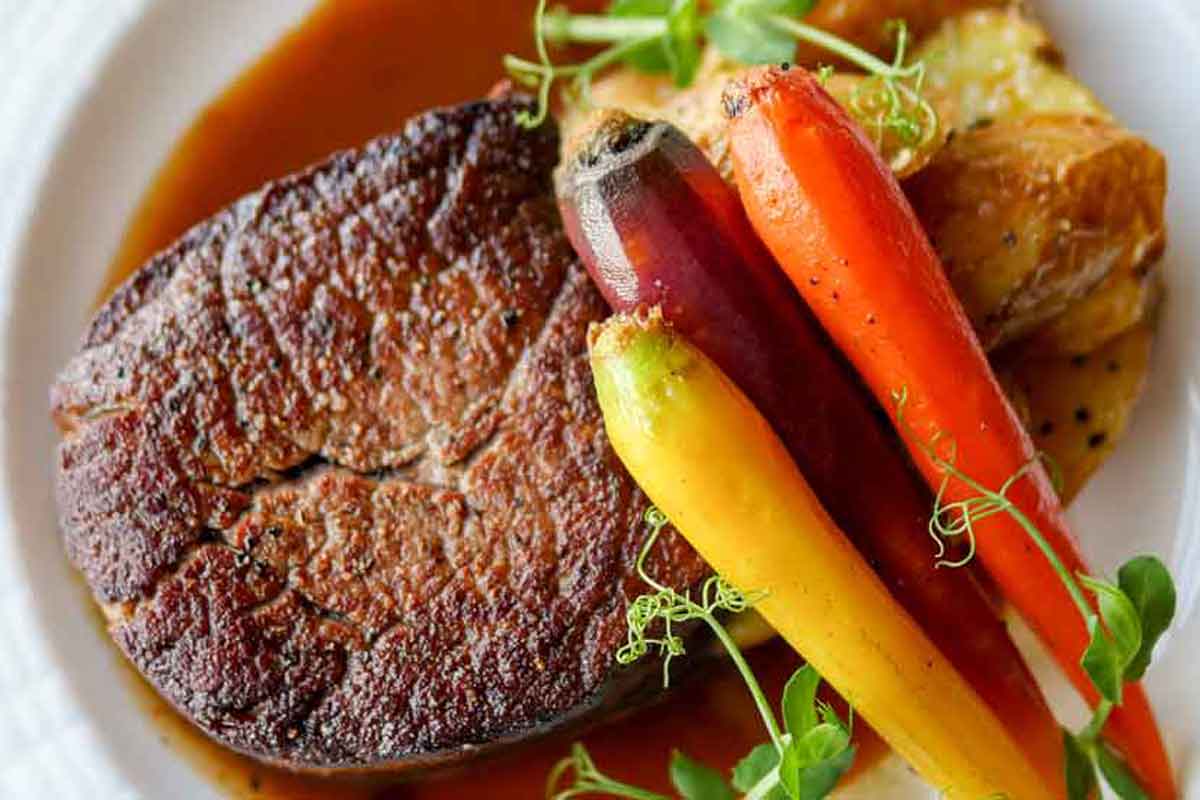 A large portion of steak with tri-color carrots and potatoes and a herb garnish with beef au jous